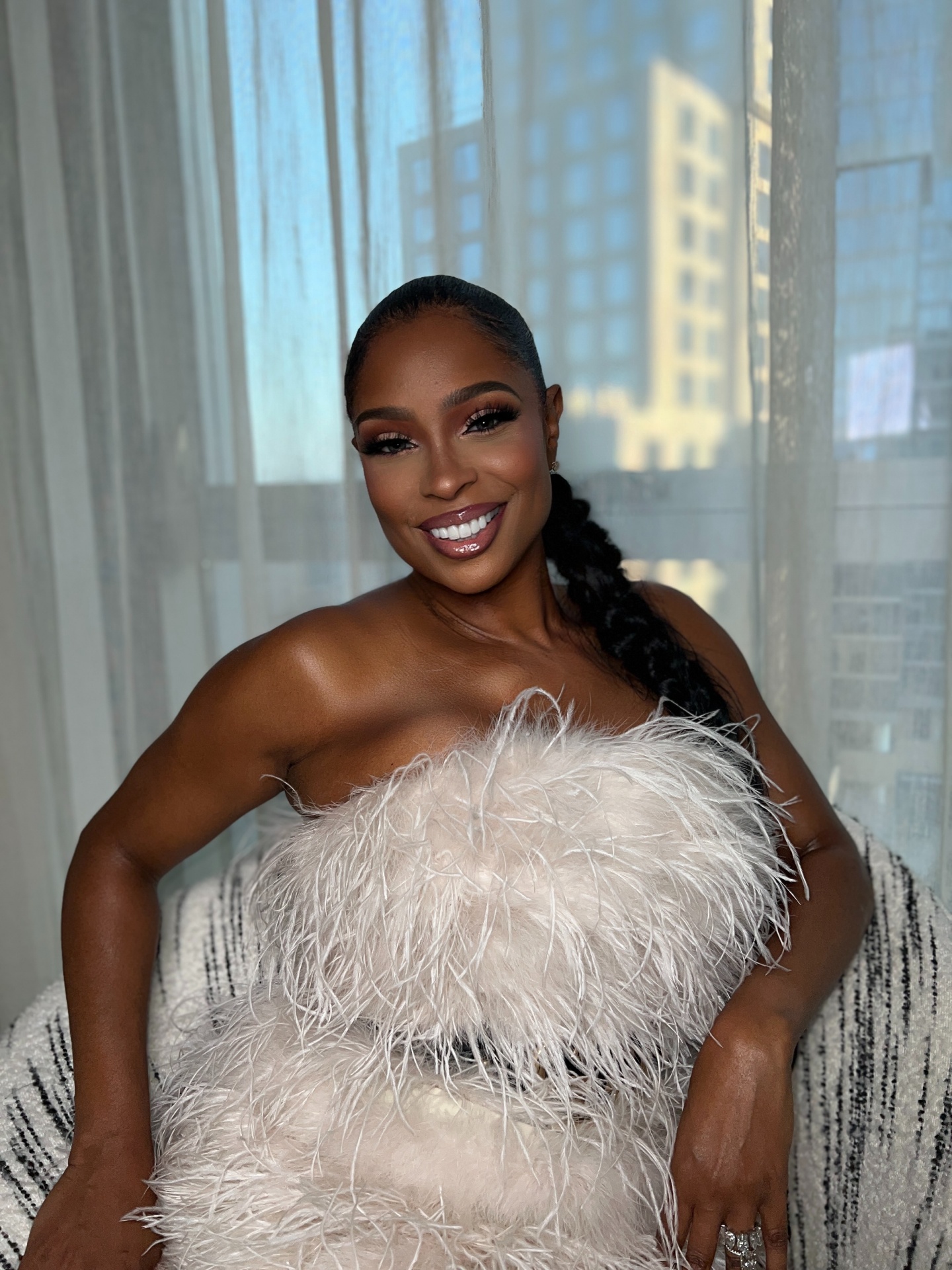 Jennifer Gold in a feathered gown seated against a city skyline.